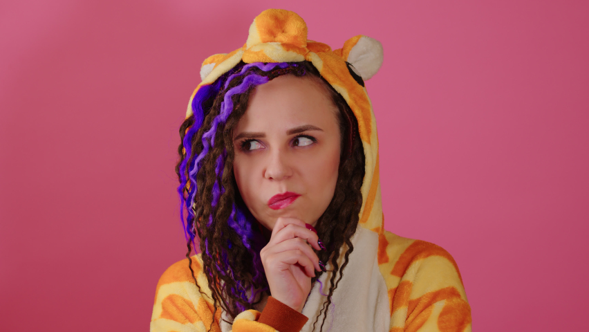 Young thoughtful woman in kigurumi scratching chin, looking around on pink background. Cute vivid female with dreadlocks in pajamas with reflecting thoughts, doubt glance. Royalty-Free Stock Footage #1090742083