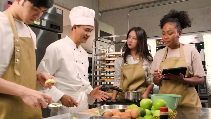 Hobby cuisine course, senior male chef in cook uniform teaches young cooking class students to peel and chop apples, ingredients for pastry foods, fruit pies in restaurant stainless steel kitchen. Royalty-Free Stock Footage #1090743727
