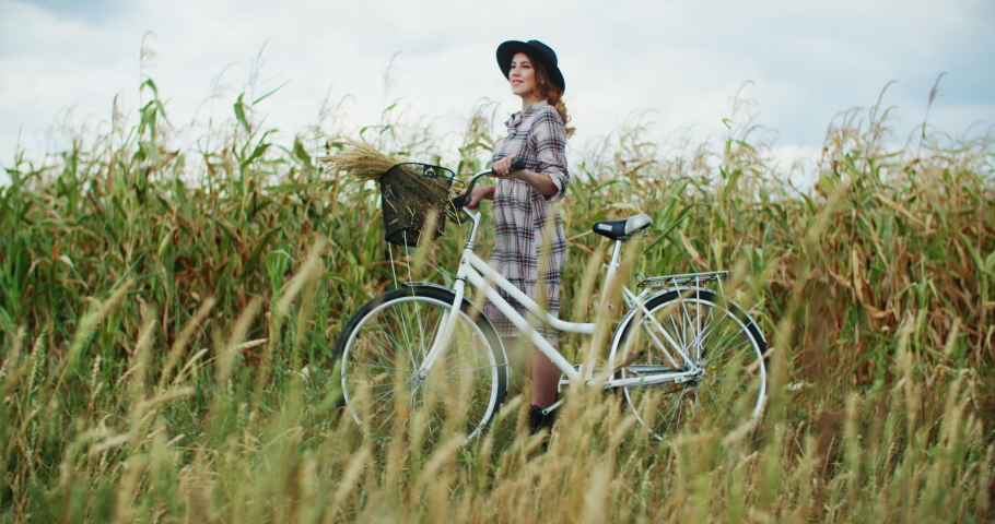 Beautiful woman in rustic check dress and black hat walking with white bike through harvest field against blue and white cloudy sky and enjoy scenery
