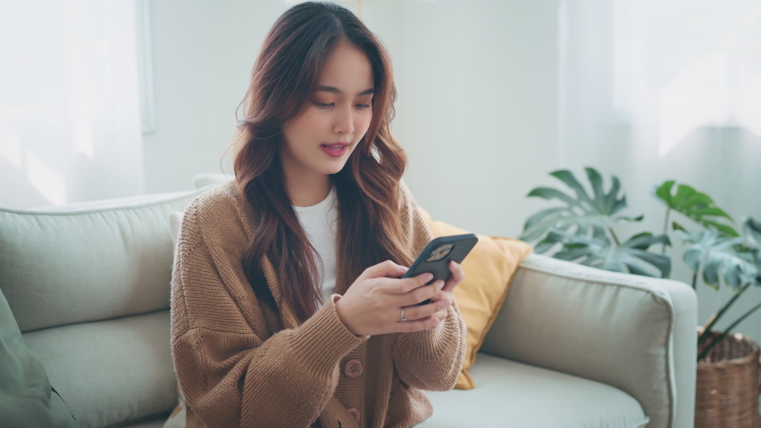 Excited young asian woman hold phone chatting in dating app feel happy sit on sofa at home, Closeup joyful female reading good news on phone in slow motion. Surprised lady celebrating victory on phone Royalty-Free Stock Footage #1090746115
