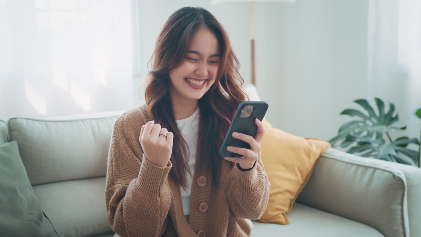 Excited young asian woman hold phone chatting in dating app feel happy sit on sofa at home, Closeup joyful female reading good news on phone in slow motion. Surprised lady celebrating victory on phone | Shutterstock HD Video #1090746115