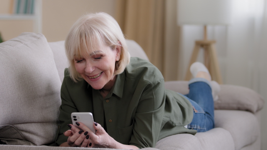 Old happy Caucasian grandmother 60 years senior woman 50s lady blonde elderly female lies on couch at home enjoys remote mobile conversation video call conference chat smiling talking to phone camera