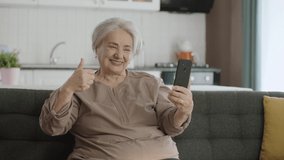 Senior woman smiling, enjoying video call with relatives, looking at camera and waving, sitting on comfortable sofa. Technology use concept with the elderly. Taking selfie with thumbs up sign.