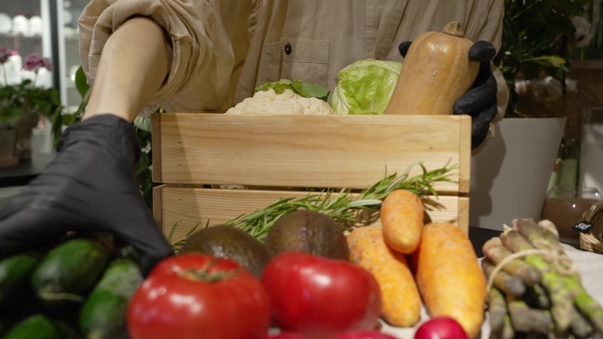 Shop worker carefully loads wooden box with variety of fresh vegetables. Woman puts sorted organic vegetables into wooden box closeup