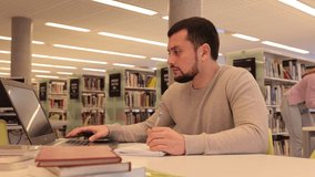 Focused guy student preparing for an exam on a laptop in the university library, looking for sources of information in textbooks. High quality 4k footage