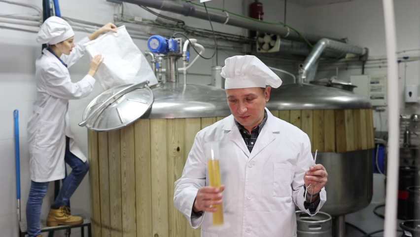 Interested brewery owner checking alcoholic content in craft beer, using test tube and hydrometer while standing near fermenters in workshop
