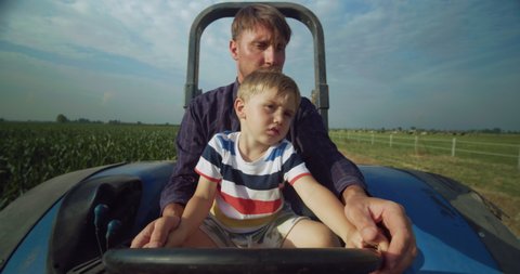Authentic shot of happy farmer father is driving a tractor with his little son and showing a family agricultural possession in a sunny day. 