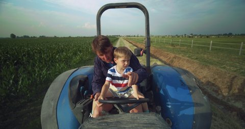 Authentic shot of happy farmer father is driving a tractor with his little son and showing a family agricultural possession in a sunny day. 