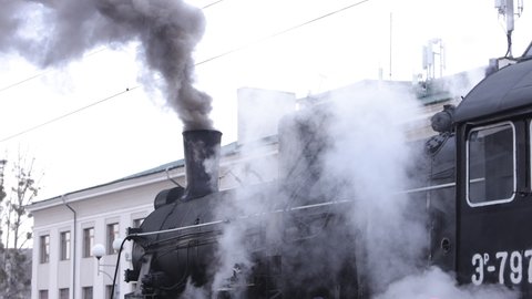 Vintage black steam locomotive. Historic train runs through fields. Vehicle departs from railway station. Old steam train locomotive is leaving the railway station.