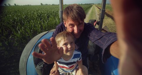 Cinematic shot of happy farmer father is having fun to make video call to family or friends together with his little son in tractor while showing a family agricultural possession in sunny day.