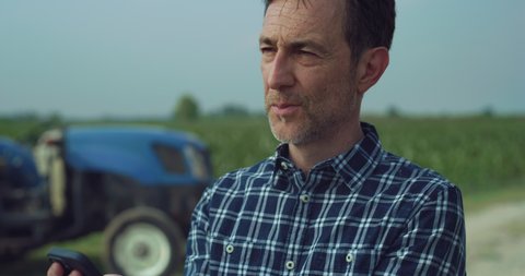 Cinematic shot of mature farmer is using smartphone and smiling satisfied with his work on countryside farmland corn fields. 