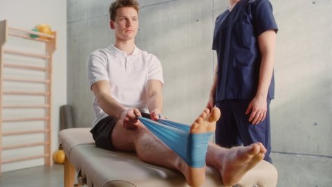 Young Male Athlete Undergoing Physiotherapy, Professional Sport Masseur Helping with Foot Exercise with a Rubber Band. Musculoskeletal Pain Therapy and Rehabilitation Concept.