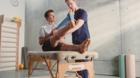Professional Sport Physiotherapist Showing Rehabilitation Exercise for Young Male Athlete's Foot with Rubber Band. Sportsman Recovering from Mild Injury. Trauma Prevention Therapy or Rehabilitation.