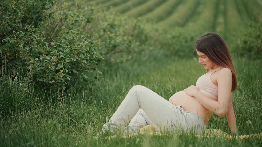 Pregnant young woman with dark hair in pink top and light pants sitting on green grass in field, woman smiling and stroking her belly with hand.