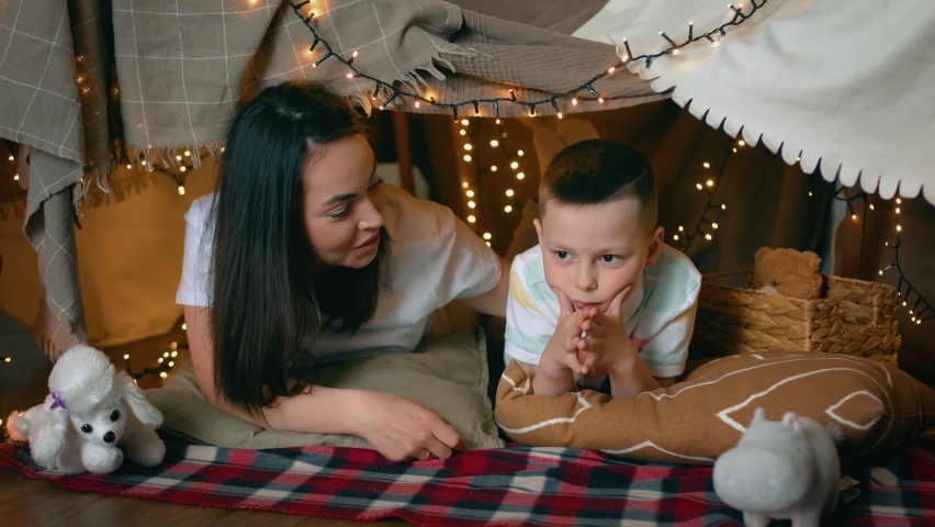 Mom and little son have fun chatting while lying in a tent of blankets and pillows in the bedroom. A family