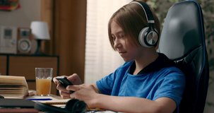 A teenager listens to music on wireless headphones while doing homework. A young boy searches for a study playlist on his phone and uses an app with songs.