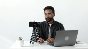 Medium shot of handsome african-american man shooting on phone camera tripod sitting at the white table with laptop and green, talking into camera during record new vlog for his internet followers 4k