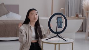 Lifestyle blogging. Young emotional asian lady influencer recording video on smartphone for social media, telling story to subscribers, sitting against circular lamp at home, slow motion