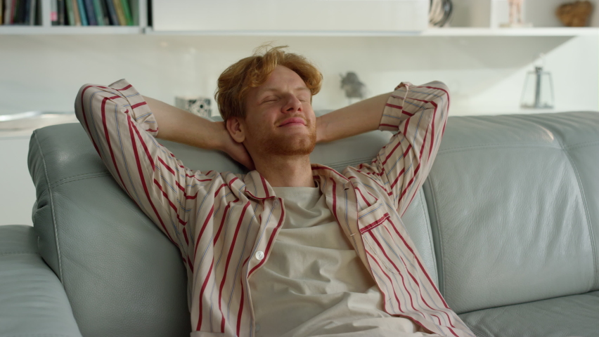 Smiling guy lean couch at cozy home closeup. Happy freelancer resting on couch enjoying siesta. Young ginger man having break taking nap in pajamas. Satisfied manager student relax chilling on weekend