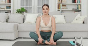Young athletic woman sitting on a mat at home in the living room preparing for sports, she looking at the video camera lens and smiling. Fitness. Concept for an active and healthy lifestyle.