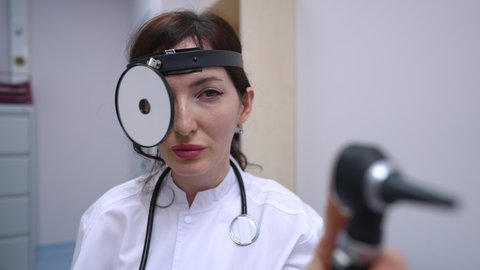 Portrait of positive Caucasian female otolaryngologist posing in slow motion with medical equipment. Beautiful woman looking at camera smiling sitting at workplace in hospital indoors