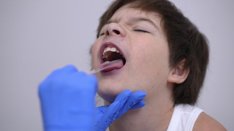 Close-up face of ill Caucasian boy opening mouth as unrecognizable doctor checking oral cavity with spatula. Professional pediatrician otolaryngologist doing diagnostics of patient in hospital indoors