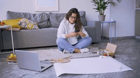 Happy young woman artist with glasses texts message on phone near large paper sheet and laptop on floor in spacious living room before painting lesson
