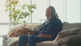 An Elderly Man with Headphones is Sitting on a Sofa with His Corgi Dog. Communication of an Elderly Owner with a Dog. The Owner Petting The Dog for His Good Behavior. Pet and Music Lover concept. - Powered by Shutterstock - Get 15% off with code: PIKWIZARD15