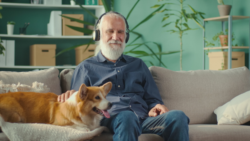 Full of Life Elderly Man with Headphones is Sitting on a Sofa with His Lovely Pet. Communication of an Elderly Owner with a Dog. Energetic Man is Enjoying the Music. Happy Life,Pet and Music Lover | Shutterstock HD Video #1090777159
