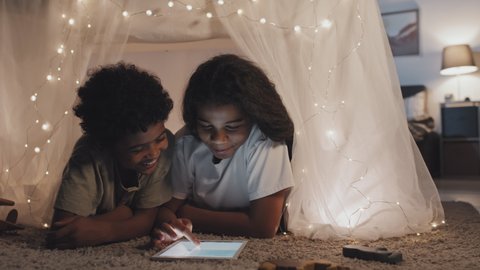 Chest-up of adorable black sister and brother lying on floor in handmade tent with lit garland at home in evening, using tablet computer, smiling and talking