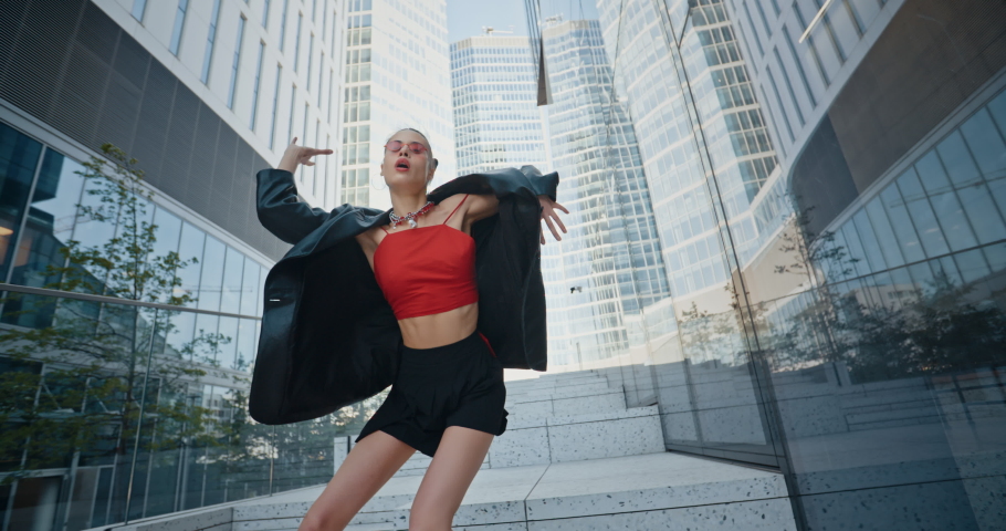 Female model wearing black jacket short skirt and cool red eye glasses dancing with hands near high rise skyscrapers in downtown district. Creative urban style woman modern city background