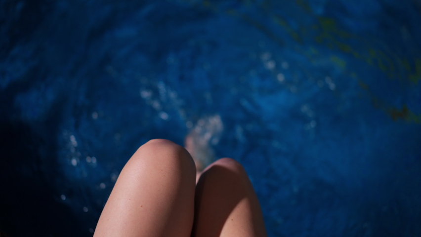 Personal POV of slim young woman splashing blue pool water in slow motion with legs. Top view waters in hotel spa with Caucasian unrecognizable tourist enjoying leisure on vacations
