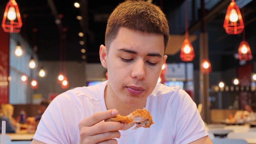 Attractive young man student in white t shirt eating chicken wings, high calorie food, health risks, cholesterol. Close up of male mouth eating junk food indoors. The guy bites a crispy chicken. | Shutterstock HD Video #1090790863