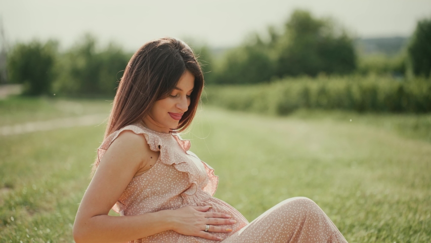 Happy young pregnant woman in a pink dress sitting among green plants and wildflowers. She gently strokes her stomach with her hands and looks at the camera with a smile.
