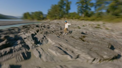 FPV freestyle drone shot active travel woman walking on sharp cliff stone structure near river water flow sunny mountain landscape. Aerial around view independent female enjoy freedom sunset scenery