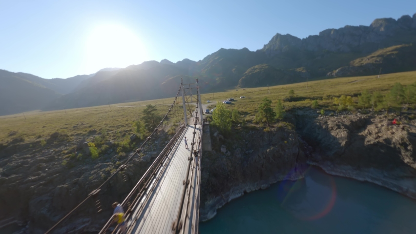 Aerial rotation view extreme couple photographing on top altitude walking bridge over mountain river at sunny landscape. FPV sports drone shot epic nature scenery cliff terrain sunset creek water flow