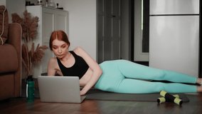 A girl in sportswear, lying on a mat, performs exercises. The girl is watching a video lesson on the laptop on how to properly perform the exercises. Sport