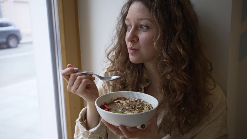 Young curly woman eats sweet vegan cereal bowl sitting on windowsill. Lady eats vegan fruit bowl with yogurt and cocoa for breakfast near window