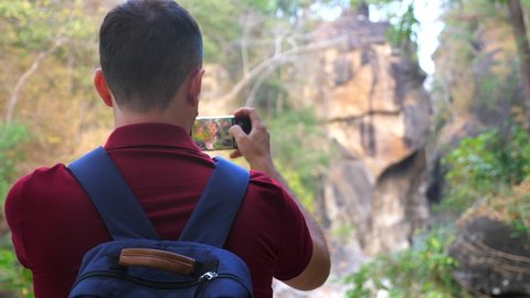 Mountain hiking photographer or video blogger man filming a canyon on cell phone. Guy traveler with a backpack videographer. Capturing scenery and nature with a smartphone panoramic camera. Back view