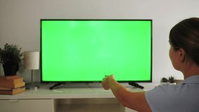 Woman watching TV with green screen switching channels, using remote control in living room.