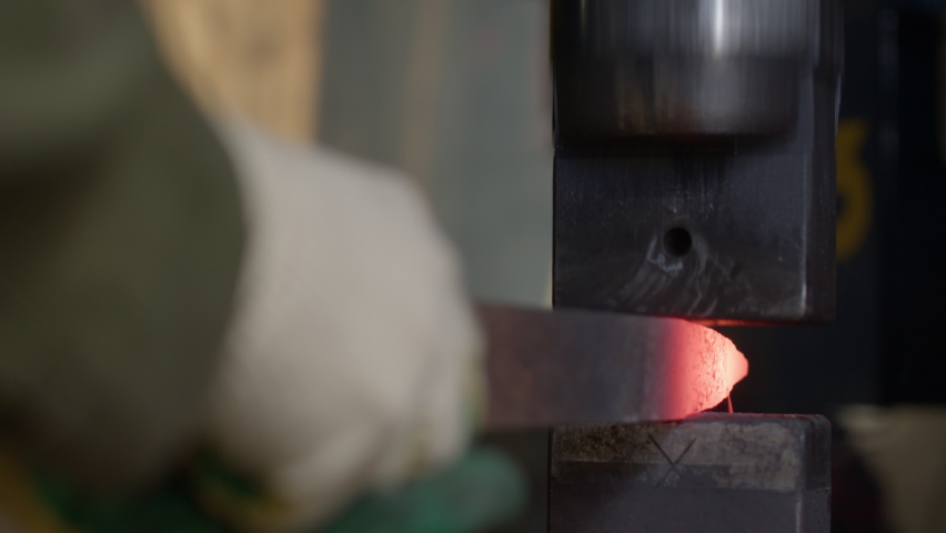 The blacksmith takes out a billet of red-hot metal from a gas furnace. A worker makes metal works of art. A mechanical anvil flattens an iron billet