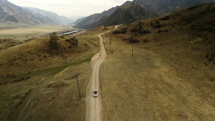 A white car drives off-road in a mountainous area. Large mountains of brown color surround the river. Aerial photography in 4k.