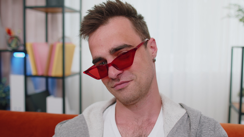 Portrait of seductive cheerful stylish successful man in t-shirt wearing red sunglasses, charming smile, blinking, winking eye, flirting. Young lovely adult guy sitting at home in living room on couch