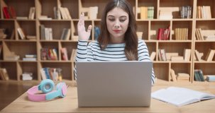 Young brunette girl in stripy shirt sits behind library desk with laptop on top and raises hand to answer, then reads from open book. Female student having online lesson in front of bookshelves.