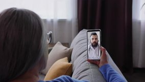 Over shoulder view of sick senior woman patient at home using smartphone for video call to her doctor, get consultation online. Video conference in medical app, video chat, telemedicine, telehealth.