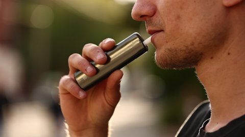 close up of a man with an electronic cigarette. man smokes an electronic cigarette in a natural background. hipster smokes an e-cigarette, breathes out streams of smoke and vaping outdoors copy space.