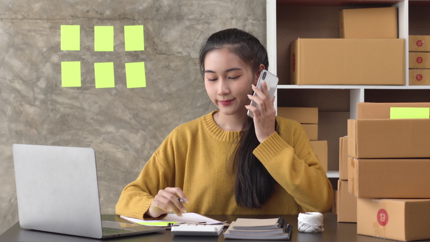 Woman entrepreneur talking with customer on a mobile phone and writing product order at home office, (SME business)