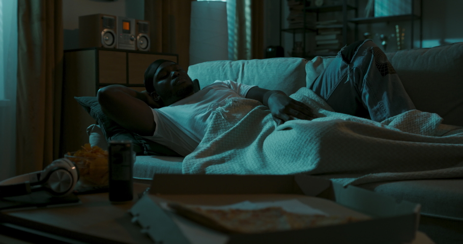 Sad, wounded young man lies in living room at night, unable to sleep, contemplating, room lit by streetlight streaming in through window. Boy is heartbroken, eating pizza and fries out of nervousness. | Shutterstock HD Video #1090841295
