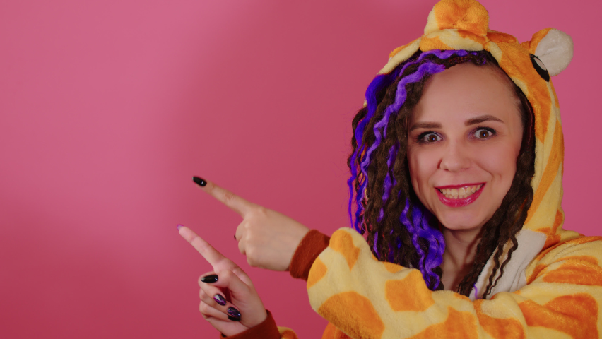 Young cute woman with dreadlocks in kigurumi pointing index fingers at space for text. Attractive female with bright curly hairstyle pointing gesture to your advertisement on pink background.