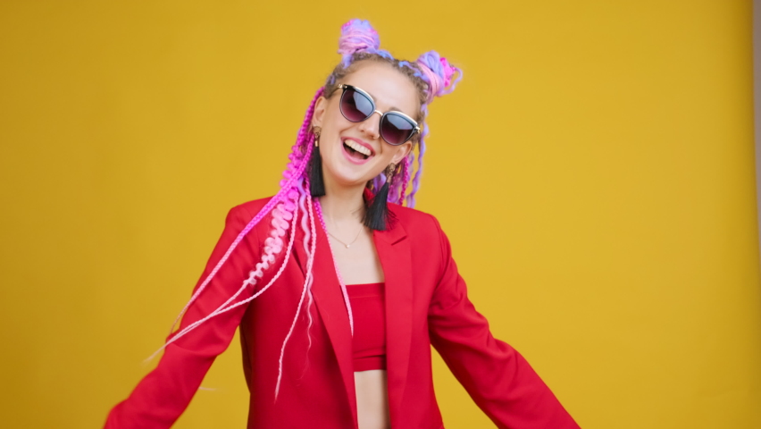 Funny young woman with original style of hair dancing in studio on blue background. She has colorful clothes, afro braids dreadlocks. Girl in sunglasses positive emotions, ecstatic feeling Royalty-Free Stock Footage #1090861525